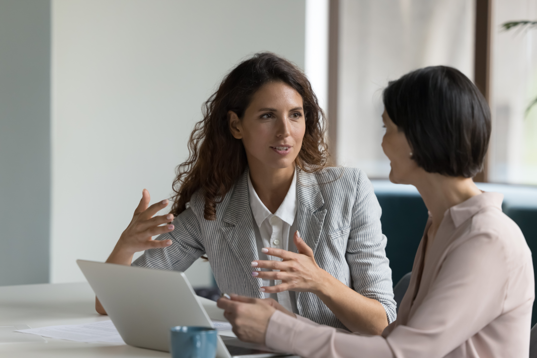 Two women, one talking with hand gestures to other using computer.