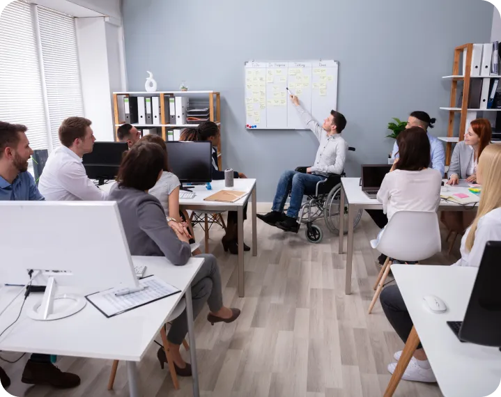 A person in a wheelchair pointing at a whiteboard with people sitting around it